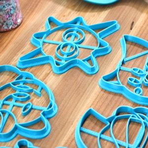 May include: Five blue plastic cookie cutters on a wooden surface. The cutters are shaped like a sun, a cat, a mouse, a bear, and a flower.