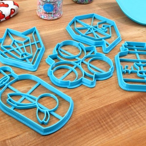 May include: Five blue plastic cookie cutters on a wooden surface. The cutters are shaped like a geometric design, a flower, a jar with the word "Honey" on it, and two bottles with a butterfly design.