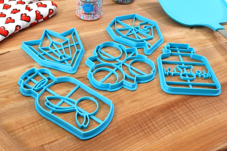May include: Five blue plastic cookie cutters on a wooden surface. The cutters are shaped like a geometric design, a flower, a jar with the word "Honey" on it, and two bottles with a butterfly design.