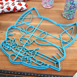 May include: A blue plastic cookie cutter shaped like a cat's face. The cutter has a detailed design with a cat's eyes, nose, and whiskers.