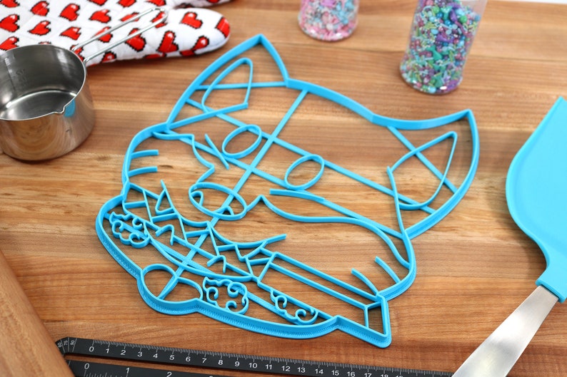 May include: A blue plastic cookie cutter shaped like a cat's face. The cutter has a detailed design with a cat's eyes, nose, and whiskers.