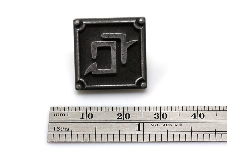 May include: A silver metal pin with a black square background and a stylized silver symbol inside. The symbol is a stylized letter "D" with a curved line extending from the top.