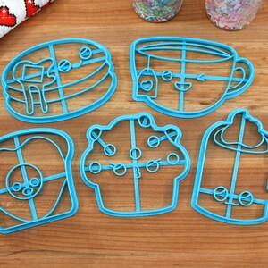May include: Five blue plastic cookie cutters shaped like a stack of pancakes, a teacup, a cupcake, a slice of toast, and a cupcake with frosting.