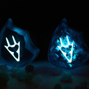 May include: Two blue resin glowing crystal shapes with a white claw mark design inside. The crystals are sitting on a dark surface with small blue and green crystals scattered around them.