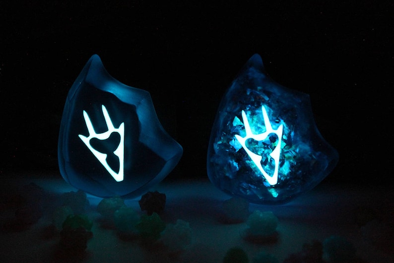 May include: Two blue resin glowing crystal shapes with a white claw mark design inside. The crystals are sitting on a dark surface with small blue and green crystals scattered around them.