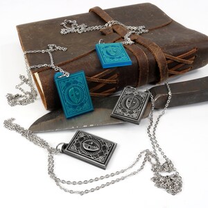 Puede incluir: Tres cadenas de plata con colgantes, cada uno con un dise&ntilde;o diferente. Dos colgantes son de plata con un dise&ntilde;o de cara en blanco y negro, y un colgante es azul con un dise&ntilde;o de cara en blanco y negro. Las cadenas est&aacute;n sobre un diario de cuero marr&oacute;n, un cuchillo de plata y un objeto de metal.