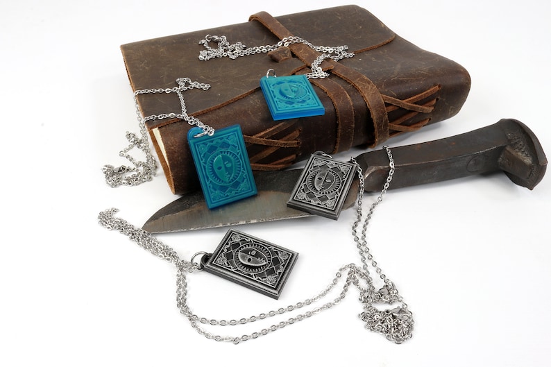 Puede incluir: Tres cadenas de plata con colgantes, cada uno con un dise&ntilde;o diferente. Dos colgantes son de plata con un dise&ntilde;o de cara en blanco y negro, y un colgante es azul con un dise&ntilde;o de cara en blanco y negro. Las cadenas est&aacute;n sobre un diario de cuero marr&oacute;n, un cuchillo de plata y un objeto de metal.