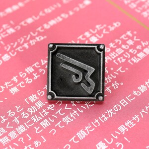 May include: A silver-toned metal pin with a stylized, abstract design. The pin is square-shaped and has a black background. The design is a stylized, abstract shape that resembles a wing or a blade. The pin is on a pink surface with Japanese text.