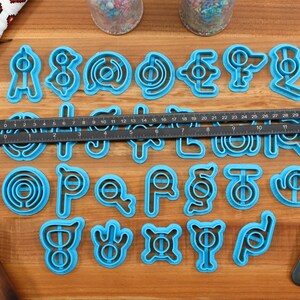 May include: A set of 26 blue plastic cookie cutters shaped like letters of the alphabet. The cutters are arranged in alphabetical order on a wooden surface.