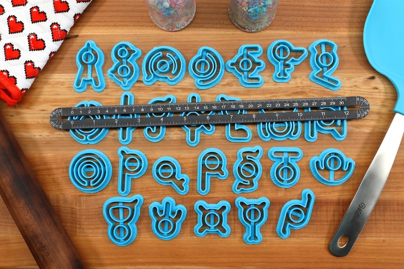 May include: A set of 26 blue plastic cookie cutters shaped like letters of the alphabet. The cutters are arranged in alphabetical order on a wooden surface.