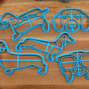 May include: Five blue plastic cookie cutters shaped like dachshunds. The cutters are on a wooden surface.