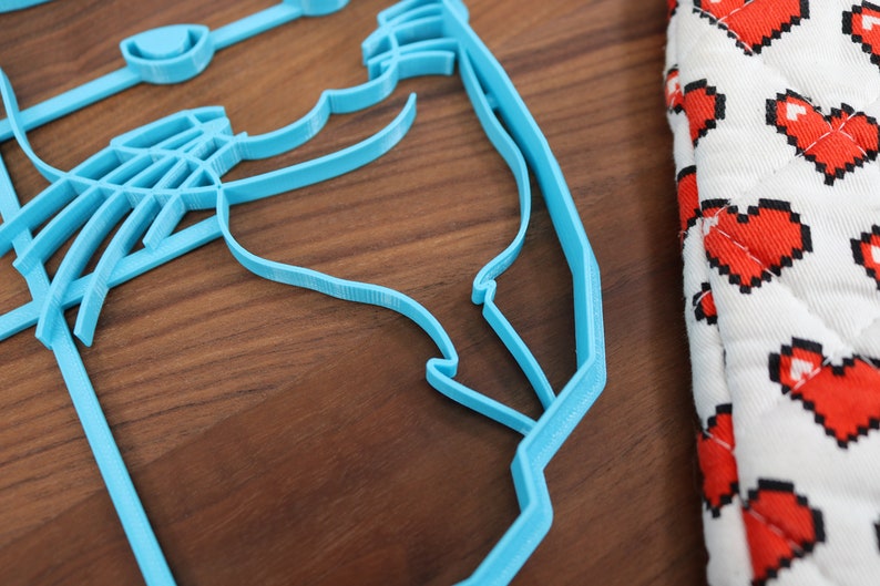 May include: A light blue plastic cookie cutter shaped like a cat's head. The cookie cutter is on a wooden surface.