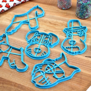 May include: Set of five blue plastic cookie cutters shaped like different variations of a schnauzer dog. The cutters are on a wooden surface.
