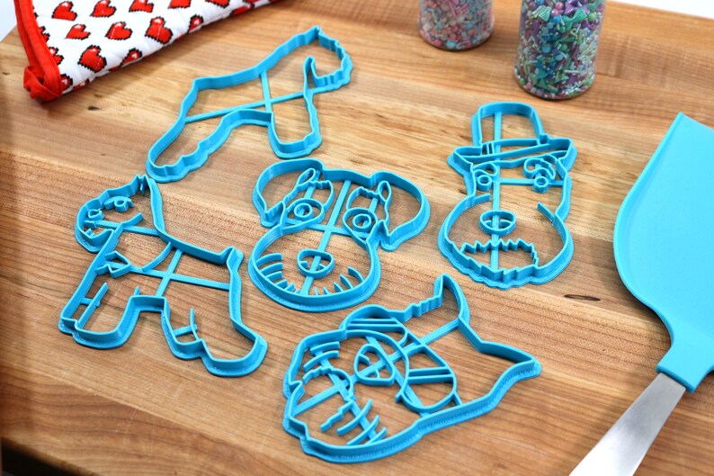 May include: Set of five blue plastic cookie cutters shaped like different variations of a schnauzer dog. The cutters are on a wooden surface.