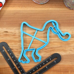 May include: A blue plastic cookie cutter shaped like a duck with its head turned to the side. The duck has a long neck and a large beak. The cookie cutter is on a wooden surface.