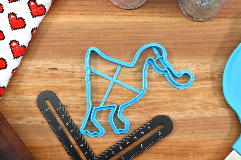 May include: A blue plastic cookie cutter shaped like a duck with its head turned to the side. The duck has a long neck and a large beak. The cookie cutter is on a wooden surface.