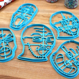 May include: Five blue plastic cookie cutters shaped like different faces. The cutters are on a wooden surface.