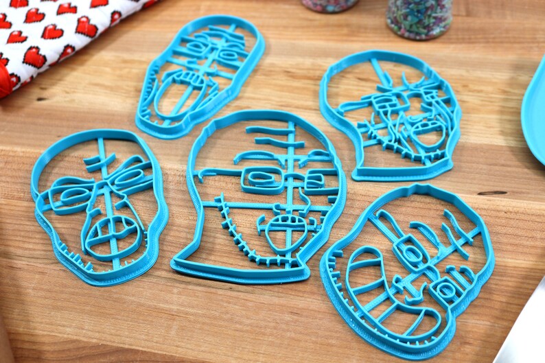 May include: Five blue plastic cookie cutters shaped like different faces. The cutters are on a wooden surface.