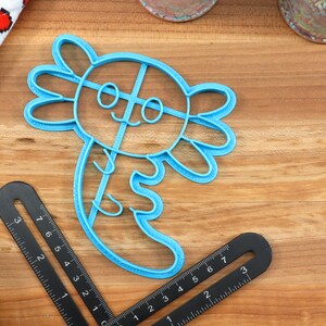 May include: A blue plastic cookie cutter shaped like an axolotl, a type of salamander. The cutter has a smiling face with two eyes and a cross-hatch pattern on its body.