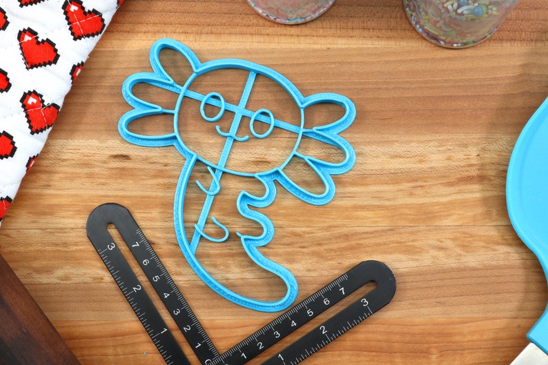 May include: A blue plastic cookie cutter shaped like an axolotl, a type of salamander. The cutter has a smiling face with two eyes and a cross-hatch pattern on its body.