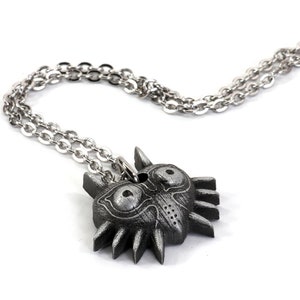 May include: A silver chain necklace with a dark gray Majora's Mask pendant. The pendant is detailed with a textured surface and features two large eyes.