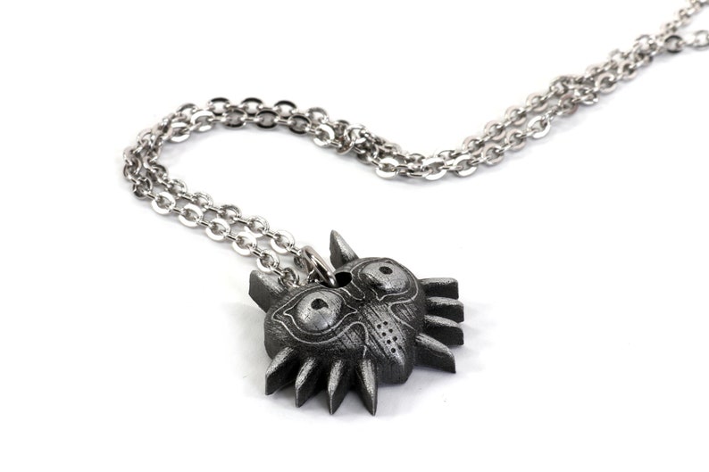 May include: A silver chain necklace with a dark gray Majora's Mask pendant. The pendant is detailed with a textured surface and features two large eyes.