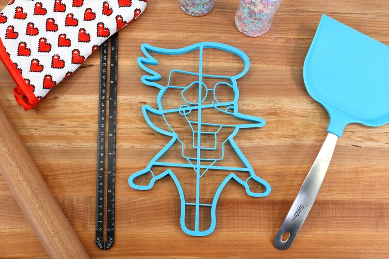 May include: A blue plastic cookie cutter shaped like a cartoon character wearing glasses. The cutter has a grid pattern inside.