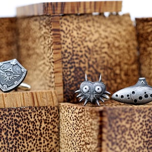 May include: Three silver cufflinks featuring iconic video game characters: a shield with a winged design, a mask with a heart shape and spikes, and an ocarina with a triangular design.