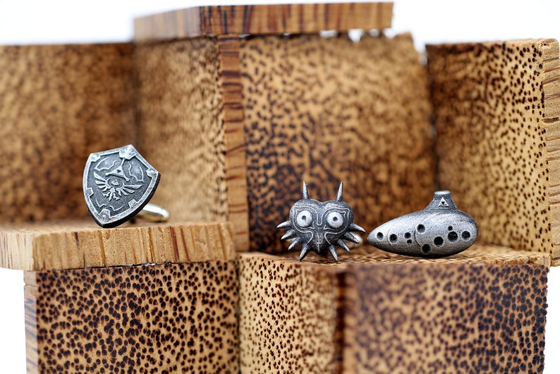 May include: Three silver cufflinks featuring iconic video game characters: a shield with a winged design, a mask with a heart shape and spikes, and an ocarina with a triangular design.