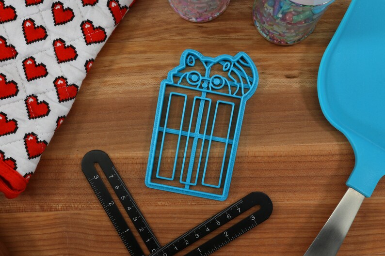 Puede incluir: Cortador de galletas de pl&aacute;stico azul con forma de mapache sosteniendo un vaso de caf&eacute; helado. El mapache tiene la nariz y los ojos negros.
