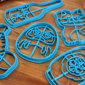 Może przedstawiać: Zestaw sześciu niebieskich plastikowych foremek do ciasteczek w kształcie butelki, kota, lalki daruma, beczki i dw&oacute;ch butelek. Formy znajdują się na drewnianej powierzchni.