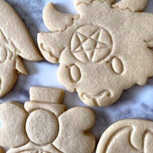 Pode incluir: Quatro biscoitos de a&ccedil;&uacute;car decorados com diferentes desenhos. Um biscoito tem um pentagrama, outro tem um rosto de desenho animado e dois t&ecirc;m desenhos florais.