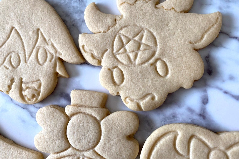 Pode incluir: Quatro biscoitos de a&ccedil;&uacute;car decorados com diferentes desenhos. Um biscoito tem um pentagrama, outro tem um rosto de desenho animado e dois t&ecirc;m desenhos florais.