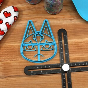 May include: A blue plastic cookie cutter shaped like a cat's face with a third eye and a sad expression. The cutter is on a wooden surface with a ruler.