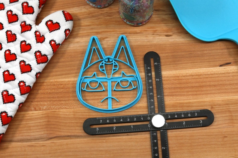May include: A blue plastic cookie cutter shaped like a cat's face with a third eye and a sad expression. The cutter is on a wooden surface with a ruler.