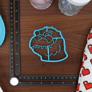 May include: Blue plastic cookie cutter shaped like a dinosaur head. The cutter has a detailed design with teeth and eyes.