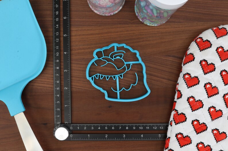 May include: Blue plastic cookie cutter shaped like a dinosaur head. The cutter has a detailed design with teeth and eyes.
