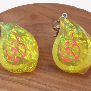 May include: Two yellow iridescent keychains shaped like teardrops with a red design inside. The keychains are on a wooden surface.