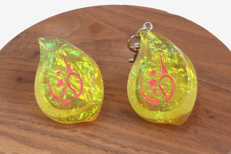 May include: Two yellow iridescent keychains shaped like teardrops with a red design inside. The keychains are on a wooden surface.