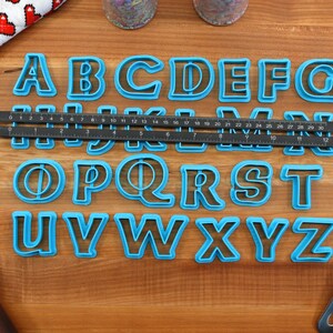 May include: A set of blue plastic alphabet cookie cutters on a wooden surface. The cutters are arranged in alphabetical order. A ruler and a spatula are also visible in the image.