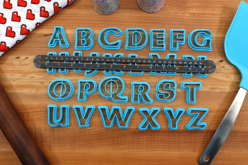 May include: A set of blue plastic alphabet cookie cutters on a wooden surface. The cutters are arranged in alphabetical order. A ruler and a spatula are also visible in the image.