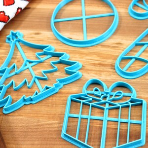 Op de afbeelding: Een set van zes blauwe plastic kerstkoekjesvormen. De vormen zijn in de vorm van een kerstboom, een snoepstok, een cadeau, een krans, een ster en een cirkel.