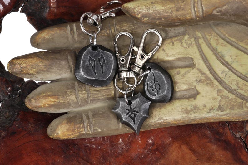 Może przedstawiać: Zestaw czterech czarnych metalowych brelok&oacute;w z r&oacute;żnymi symbolami. Breloki są przymocowane do srebrnego łańcuszka i wiszą na drewnianej dłoni.
