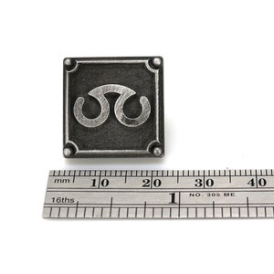 May include: A silver-colored metal pin with a square shape and a stylized symbol in the center. The pin is approximately 2 cm wide.
