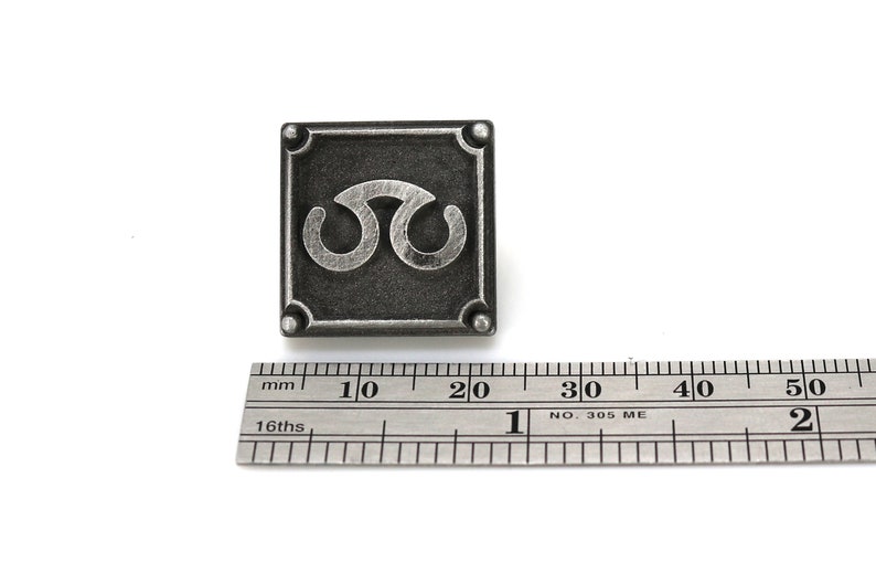 May include: A silver-colored metal pin with a square shape and a stylized symbol in the center. The pin is approximately 2 cm wide.