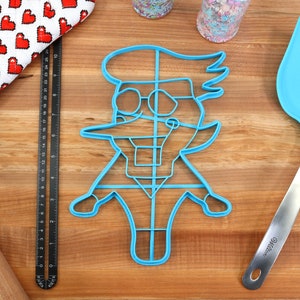 May include: Blue plastic cookie cutter shaped like a cartoon character with a large nose, glasses, and a hat. The cookie cutter is on a wooden surface.