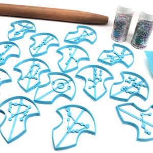 Puede incluir: Un juego de 12 cortadores de galletas de pl&aacute;stico azul con forma de constelaciones. Los cortadores est&aacute;n dispuestos sobre una superficie blanca con un rodillo, dos frascos de espolvorear y una esp&aacute;tula azul.