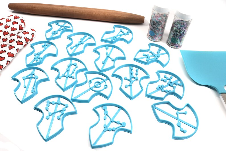 Puede incluir: Un juego de 12 cortadores de galletas de pl&aacute;stico azul con forma de constelaciones. Los cortadores est&aacute;n dispuestos sobre una superficie blanca con un rodillo, dos frascos de espolvorear y una esp&aacute;tula azul.
