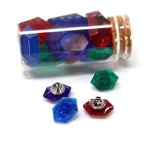 May include: A glass bottle filled with colorful plastic gems, with four gems on a white surface. The gems are red, blue, green, and purple. Each gem has a silver pin on the back.