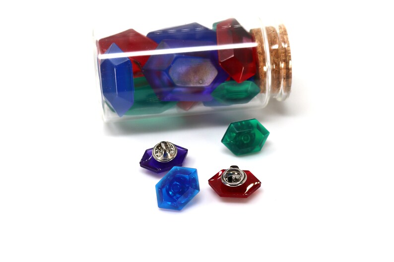 May include: A glass bottle filled with colorful plastic gems, with four gems on a white surface. The gems are red, blue, green, and purple. Each gem has a silver pin on the back.